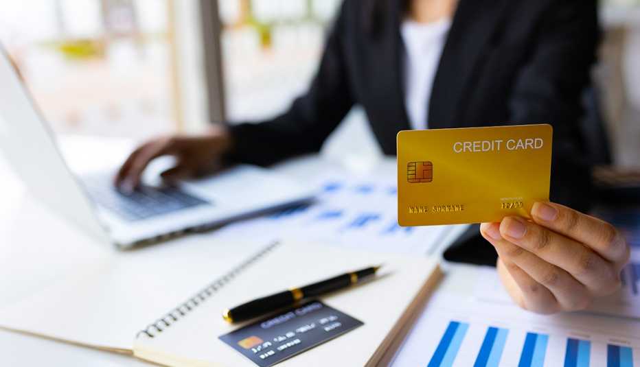  business woman holding up credit card while working on laptop computer with paperwork spread across desktop