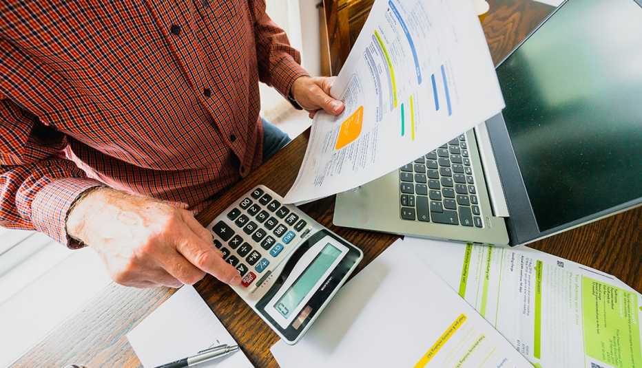 Wide angle showing a man's hand holding an energy bill while the other hand checks the numbers on a calculator. The table is strewn with documents and also a laptop.