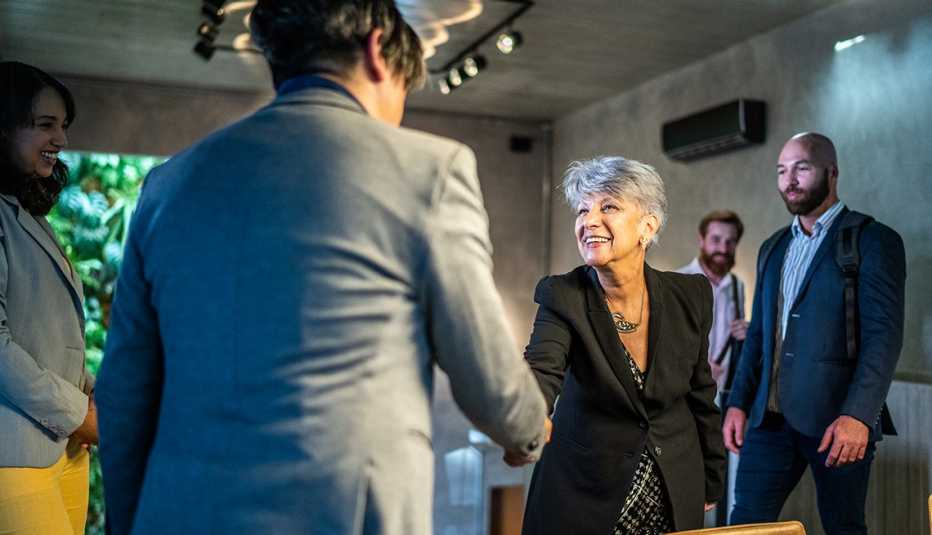 a woman in business attire shakes hands with a man in an office environment surrounded by colleagues
