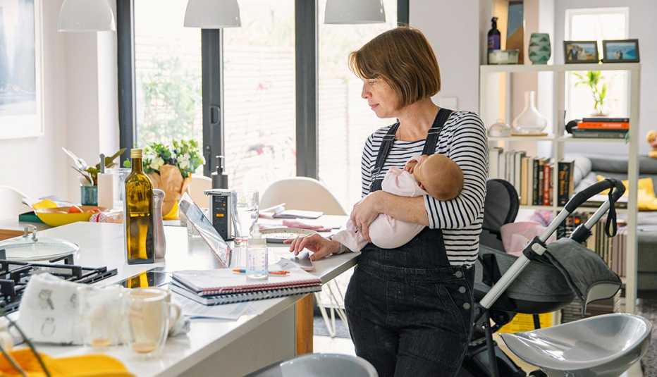 Older mother holding baby at home while working on laptop in kitchen Older mother holding baby at home while working on laptop in kitchen