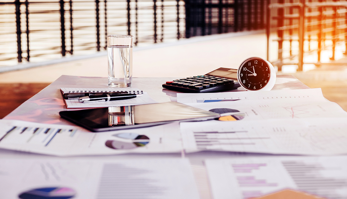 soft focus display of financial charts, a computer tablet, notebook, glass of water, calculator. and clock on a table with a view of water seen outside a window in the background