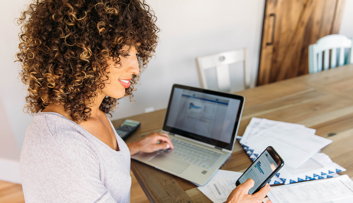 A woman seated at the dining table looks confident managing her finances with her smartphone, laptop and print out account statements