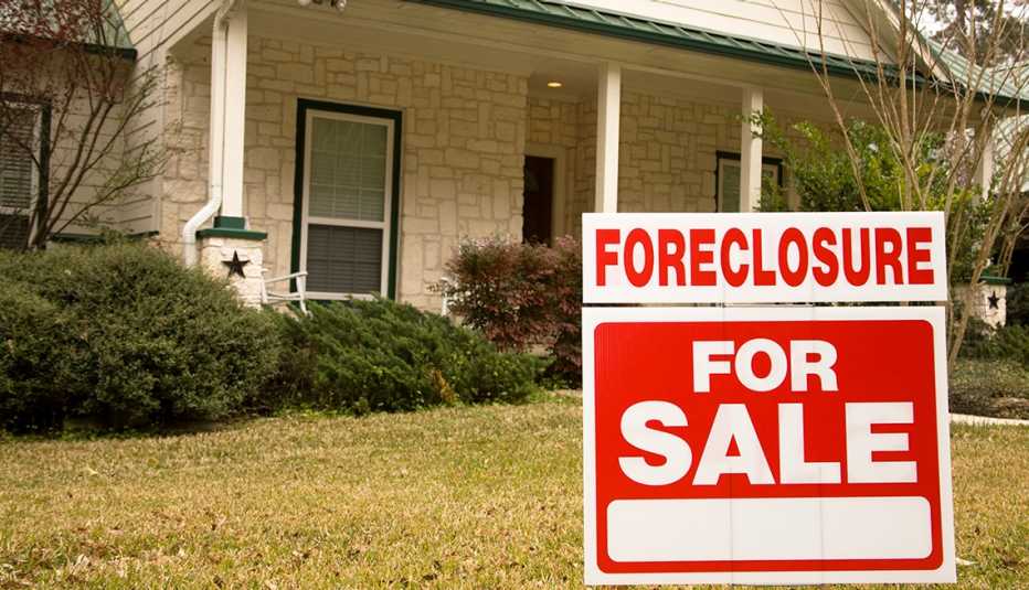 White stone home with green roof and shrubbery in front. A red and white sign in the foreground reads "Foreclosure - For Sale." White stone home with green roof and shrubbery in front. A red and white sign in the foreground reads "Foreclosure - For Sale."