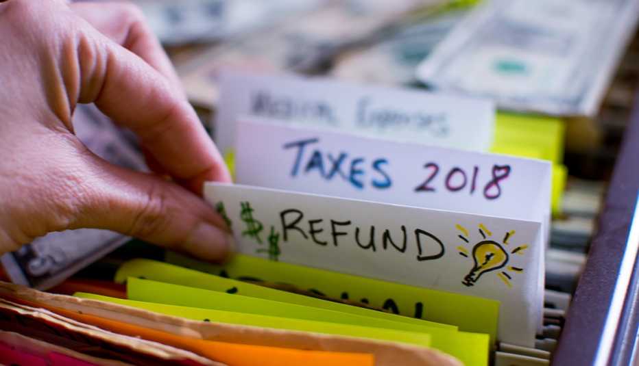Hand pulling out file in filing cabinet with tax forms and papers with words Refund and Taxes 2018 handwritten on file folder conceptual dollar signs images and light bulb for ideas on tax refund