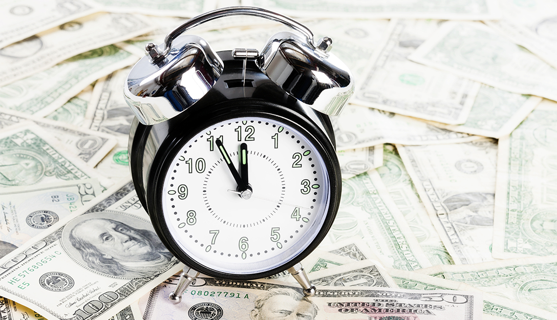 A dial alarm clock nearing midnight sits on a pile of U.S. paper currency money.