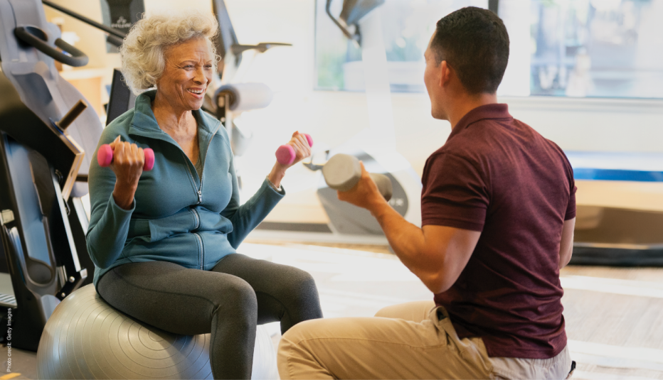 A woman sitting on an exercise ball holding small weights talking to a man sitting across from her showing her how to exercise