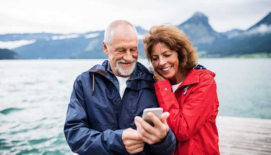 A man and a woman look at a smartphone while standing on a dock in a lake near mountains