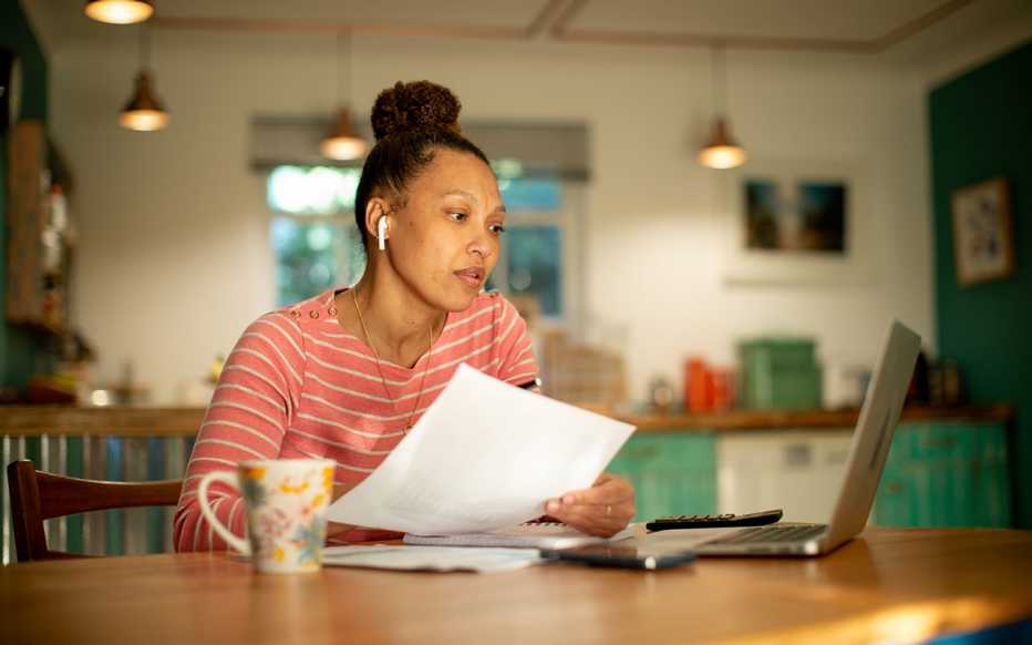 a woman sits in front of a laptop while holding a piece of paper