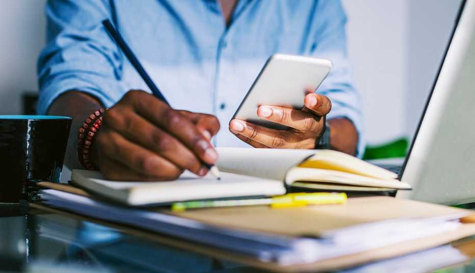 man writing in a notebook while holding mobile phone in other hand