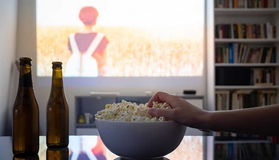 a person's hand reaches out for a bowl of popcorn next to two bottles in front of a projection on the wall a person's hand reaches out for a bowl of popcorn next to two bottles in front of a projection on the wall