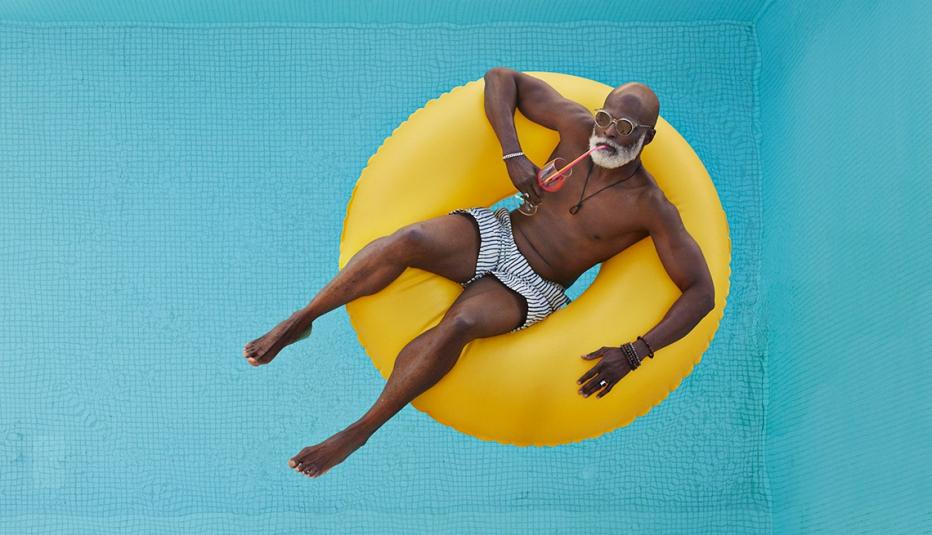 Bald senior man drinking juice while relaxing in a pool floating in a yellow inflatable ring.