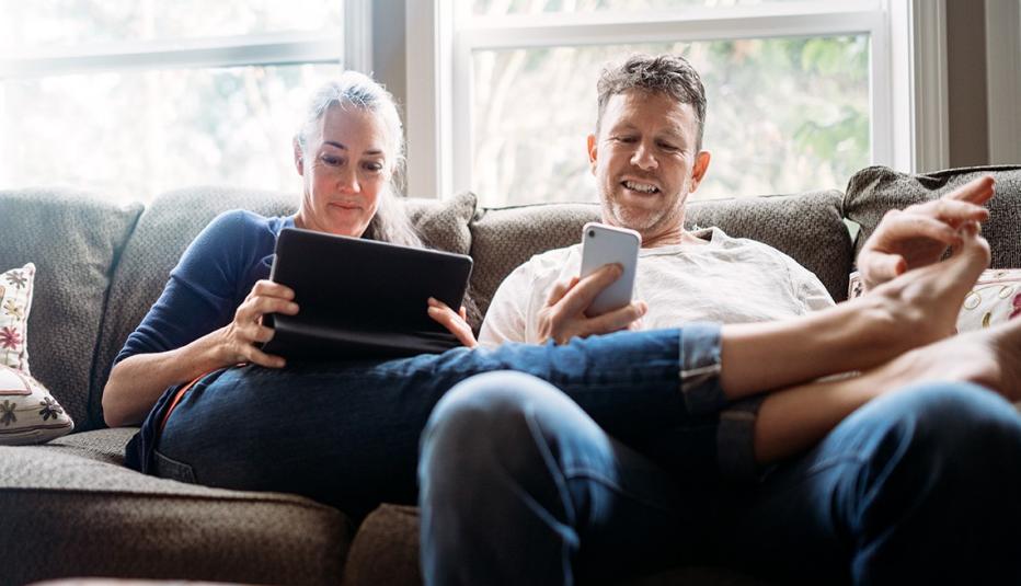 Older couple on couch relaxing and using smartphone and tablet.