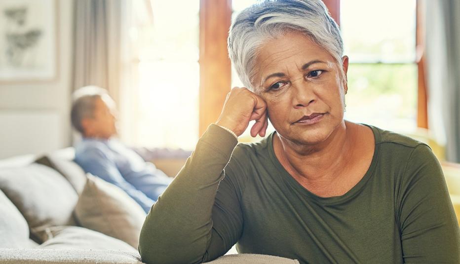 A woman sitting on her couch looking sad as she dreads her upcoming caregiving duties. A blurred image of an older man is in the background