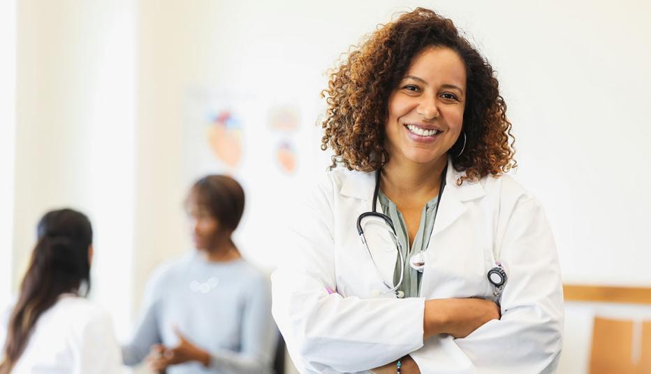 A confident female doctor smiles at the camera with her arms crossed. A colleague and patient speak in background.