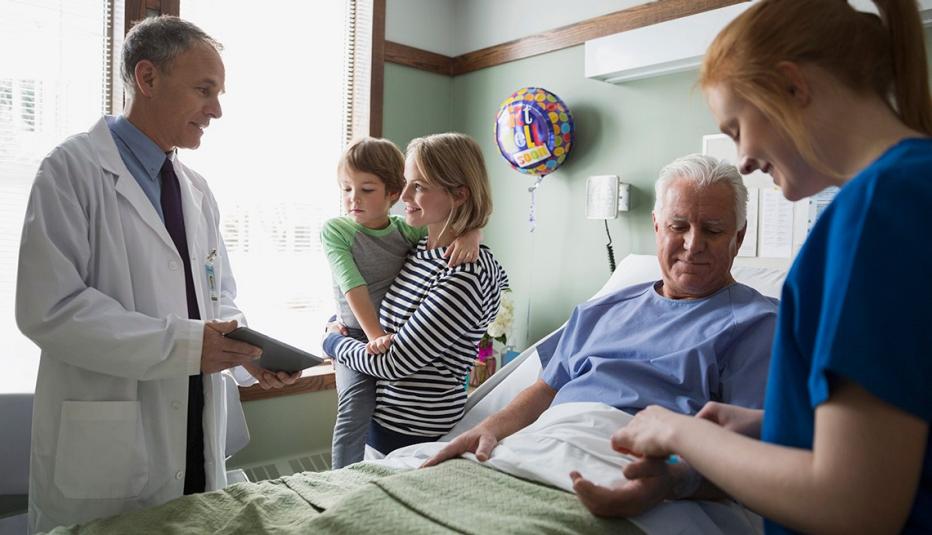 Doctor Talks To Adult Daughter Caregiver While Her Father Lays In Hospital Bed Attended To By Nurse, Handling A Hospitalization