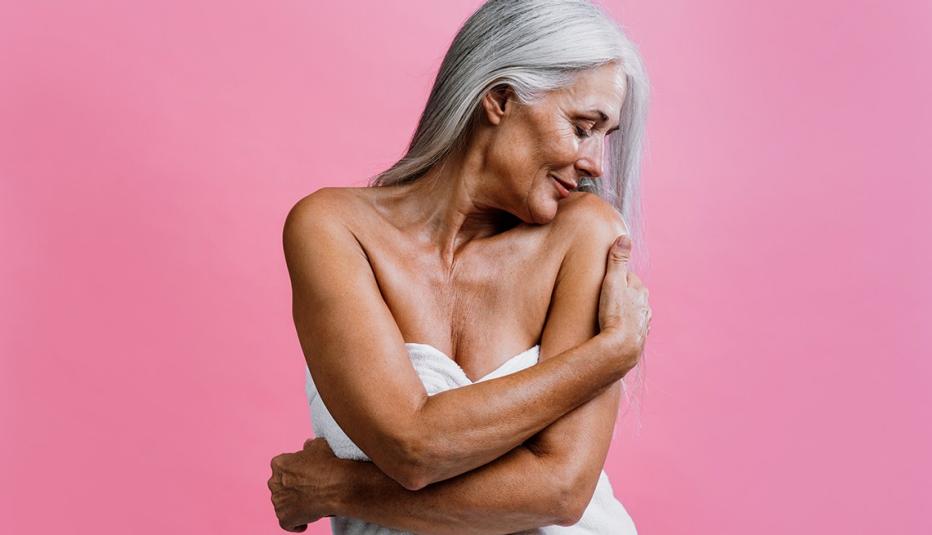 Image of a beautiful senior woman dressed in a towel posing on a pink background. Her expressions suggests body positivity, self esteem and body acceptance.