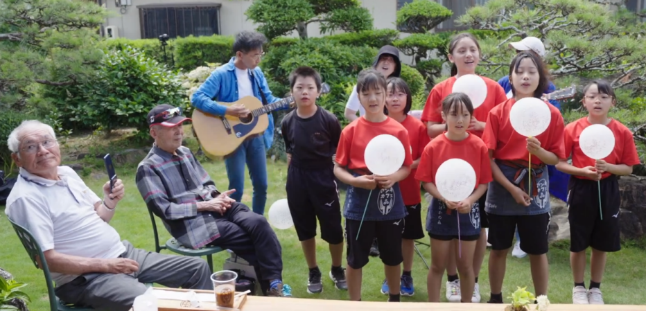 Building Empathy Across Generations at a Cafe in Japan two older men sitting outside watching group of children singing with balloons in their hands