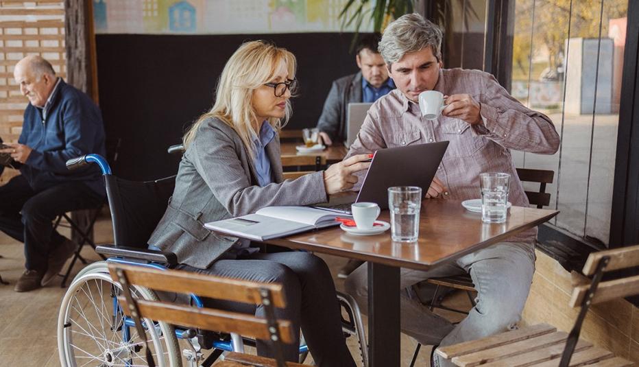 A business meeting in a coffee shop between a businesswoman using a wheelchair and a man. The woman is using laptop and note pad. Other adults can be seen sitting behind.