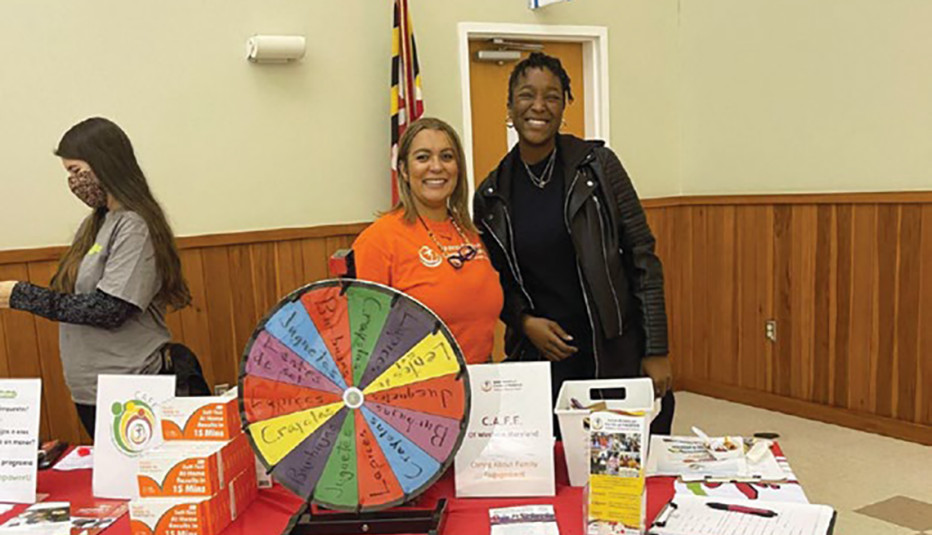 2 people standing at indoor event in back of a table with flyers and papers on it