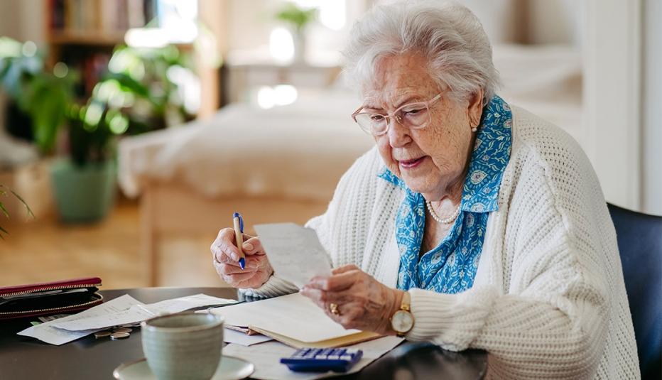 An older woman with white hair sits at her kitchen table with receipts, bills, her wallet.