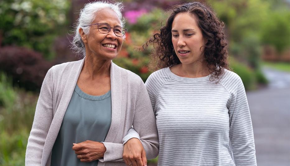 A senior woman links arms with her millennial Eurasian daughter as they happily walk through a natural parkland area and enjoy their time together.
