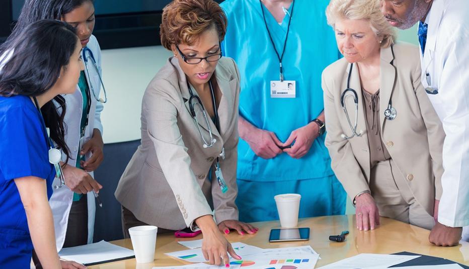 Diverse mid adult nurses, mature adult hospital adminstrators, and senior adult doctors are standing around table in hospital board room. They are reviewing spreadsheets and charts containing financial information for hospital during board meeting. Staff is wearing professional clothing, scrubs, and lab coats.