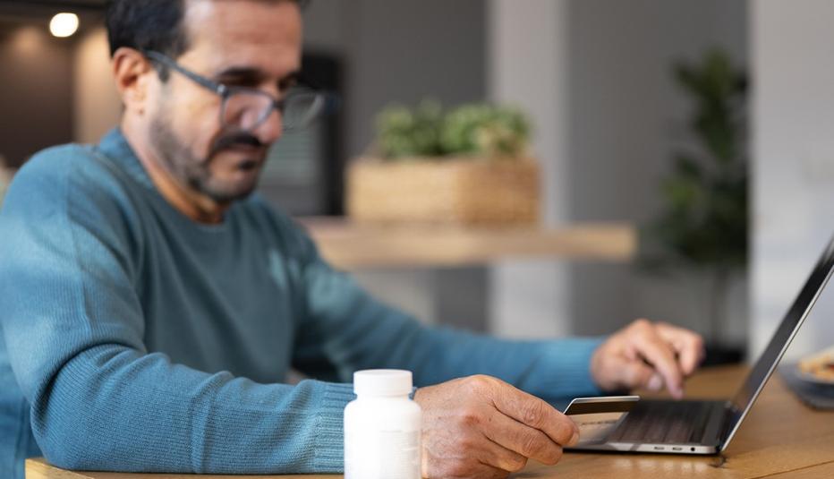 Older white man with glasses sits at kitchen table looking at his laptop with a credit card in one hand and a prescription bottle sitting next to him.