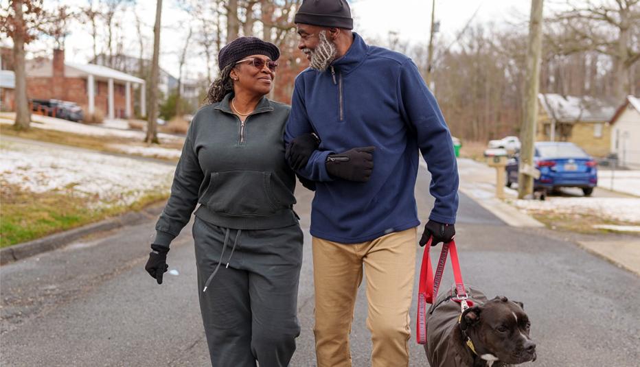 An active African American senior couple walk arm in arm and enjoy good conversation while taking their pet dog on a walk through their residential neighborhood on a cool winter day.
