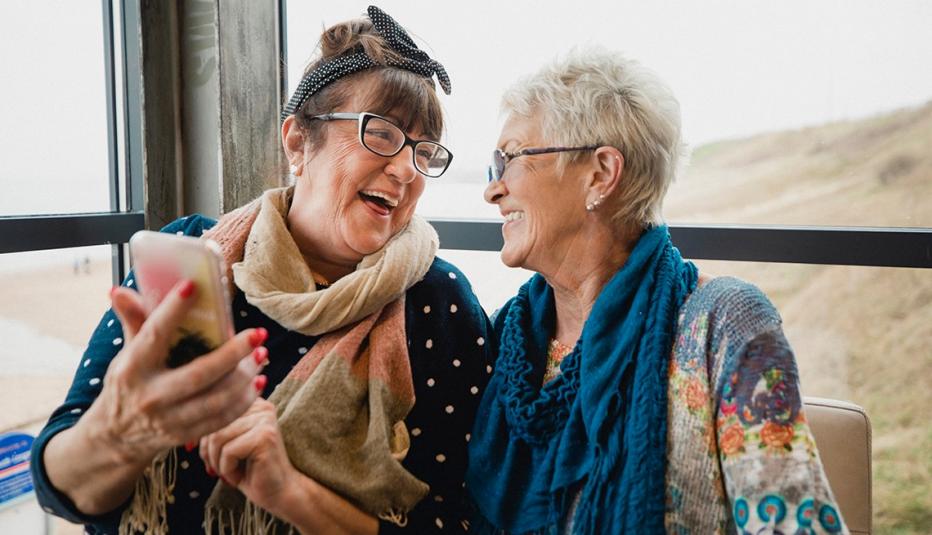 Senior female friends catching up over coffee in a seaside cafe. They are chatting to one another and sharing photos on a smartphone.
