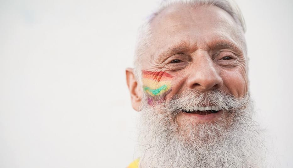 Face of a senior gay man smiling during LGBTQ+ Pride. He has a rainbow painted on his right cheek.
