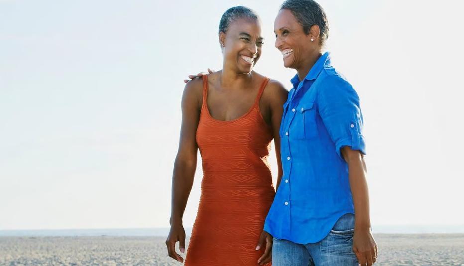 Women Walking on the Beach Two women are walking on the beach with their arms around each other's waist. One wears an orange dress; the other is wearing jeans and blue jeans.