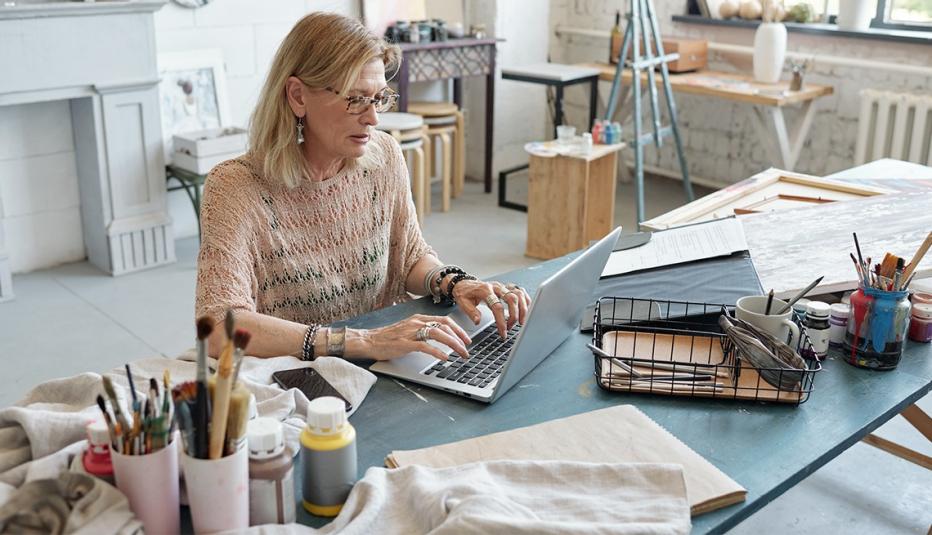 Concentrated mature artist in eyeglasses sitting at table and typing on laptop while buying consumable item for painting

