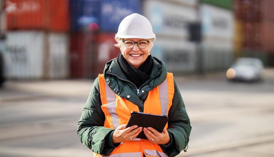 Portrait of successful female engineer with digital tablet at container warehouse. Senior woman in protective workwear looking at camera and smiling.