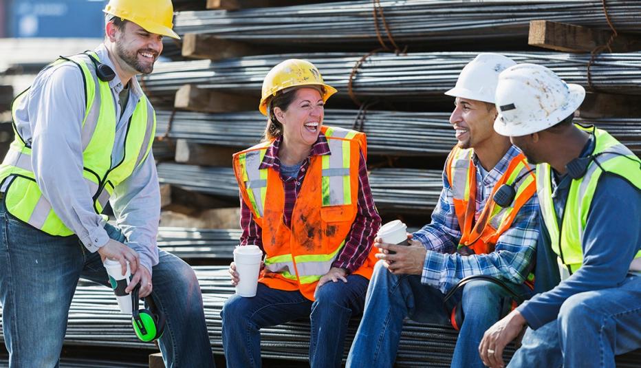 A multi-ethnic group of workers, a woman and three men, wearing safety vests and hardhats taking a coffee break. They are sitting on a stack of steel rebars (construction material), drinking coffee and talking.