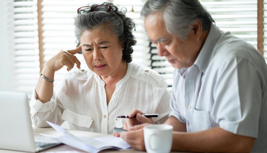 Mature Asian couple with a distressed expression examining papers in front of a laptop screen.