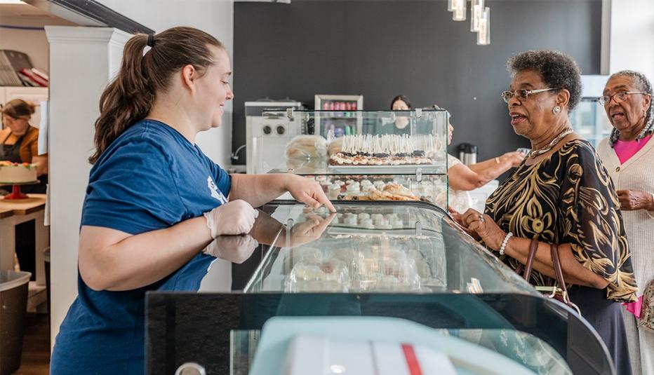 A bakery employee, dressed in a blue T-shirt and gloves, provides warm and attentive customer service to two elderly women at the counter. The bakery's clean, modern interior and neatly arranged display of baked goods create an inviting atmosphere, emphasizing the personal and friendly nature of the customer interactions