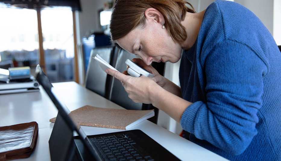 Blind woman reading document through magnifying glass while sitting next to open laptop at home
