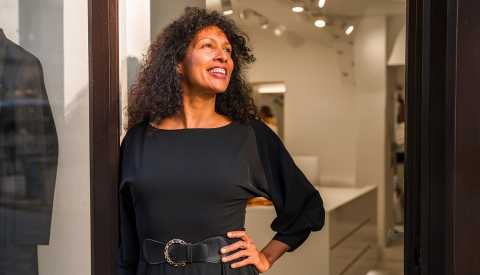 A woman posing in front of a shop window wearing a black fashionable garment with a belt, looking away and smiling, waist up image with natural sunlight