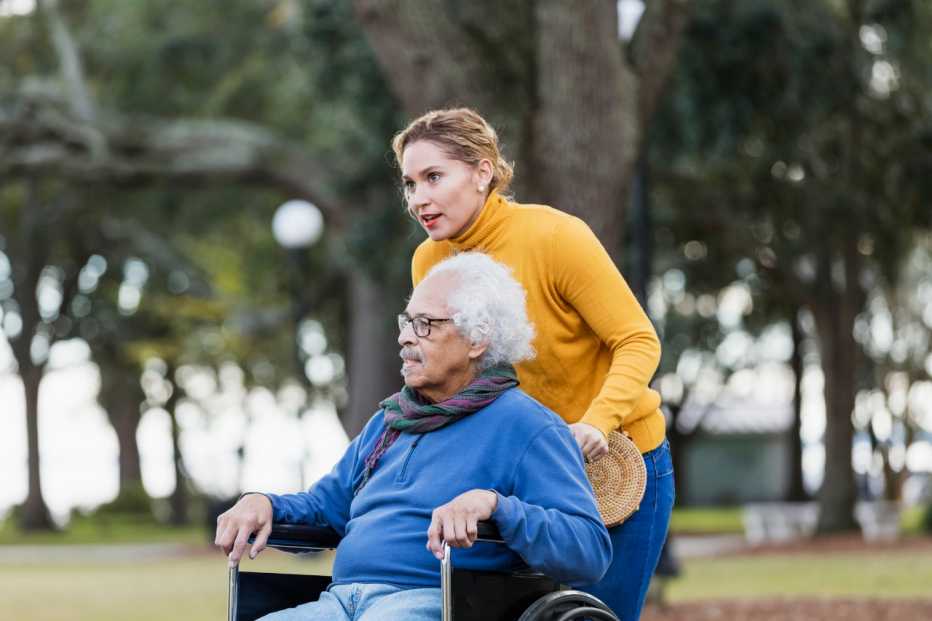 A senior Hispanic man in his 80s sitting in a wheelchair, taking a walk in the park with his adult daughter, a mid adult woman in her 30s.