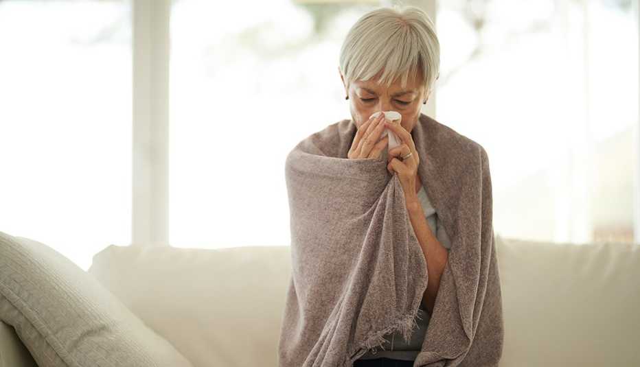 A woman on a sofa wrapped in a blanket while blowing her nose