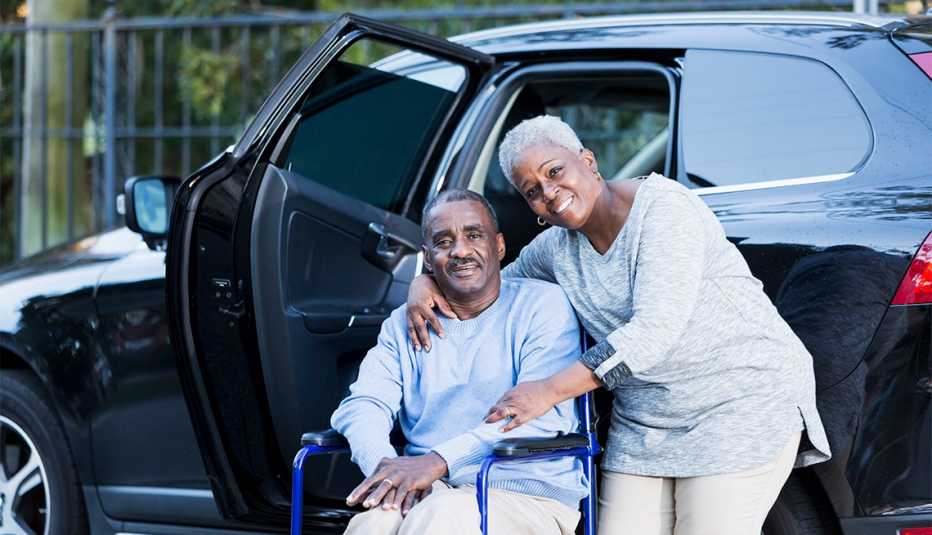 A disabled man has gotten out of a car, and is sitting in his wheelchair.  His loving wife is standing beside, with her arm around him