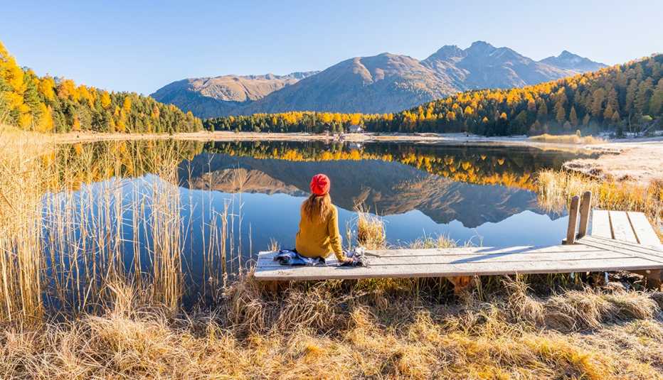 person sitting on small deck facing body of water, with trees and mountains in the distance