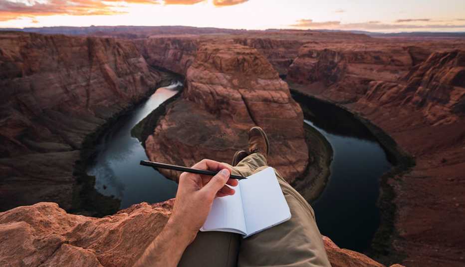 hand holding pen over notebook that is resting on person's legs; overlooking giant rock sculptures and water