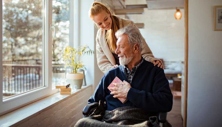 a woman puts her hands on the shoulders of a man in a wheelchair while he looks out a window
