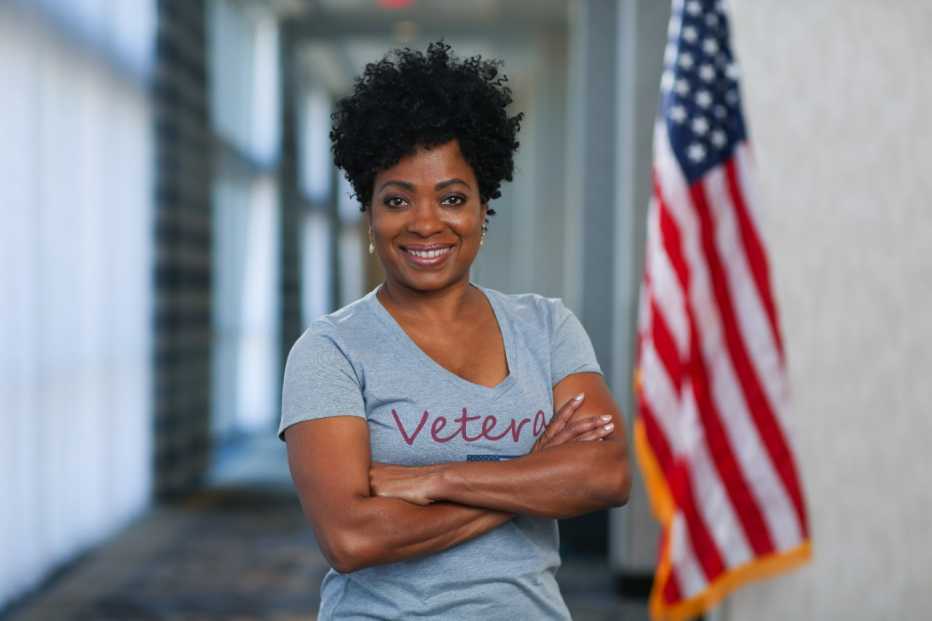 a woman standing with arms crossed in front of an american flag. her shirt has the word veteran on it