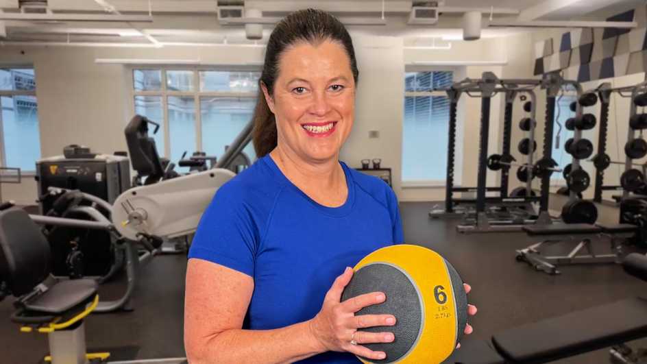 woman in a blue shirt standing in a gym while holding a medicine ball