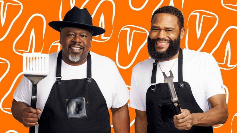 a photo shows Anthony Anderson and Cedric the Entertainer in front of a food-themed backdrop. They are wearing overalls and wielding grilling utensils