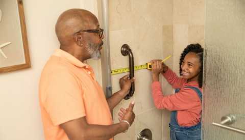 AARP HomeFit® Guide father and young daughter in shower measuring tiles  with measuring tape and pencil