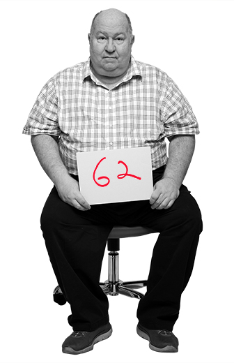 Floyd Cardwell sits in office desk chair holding placard that reads 62