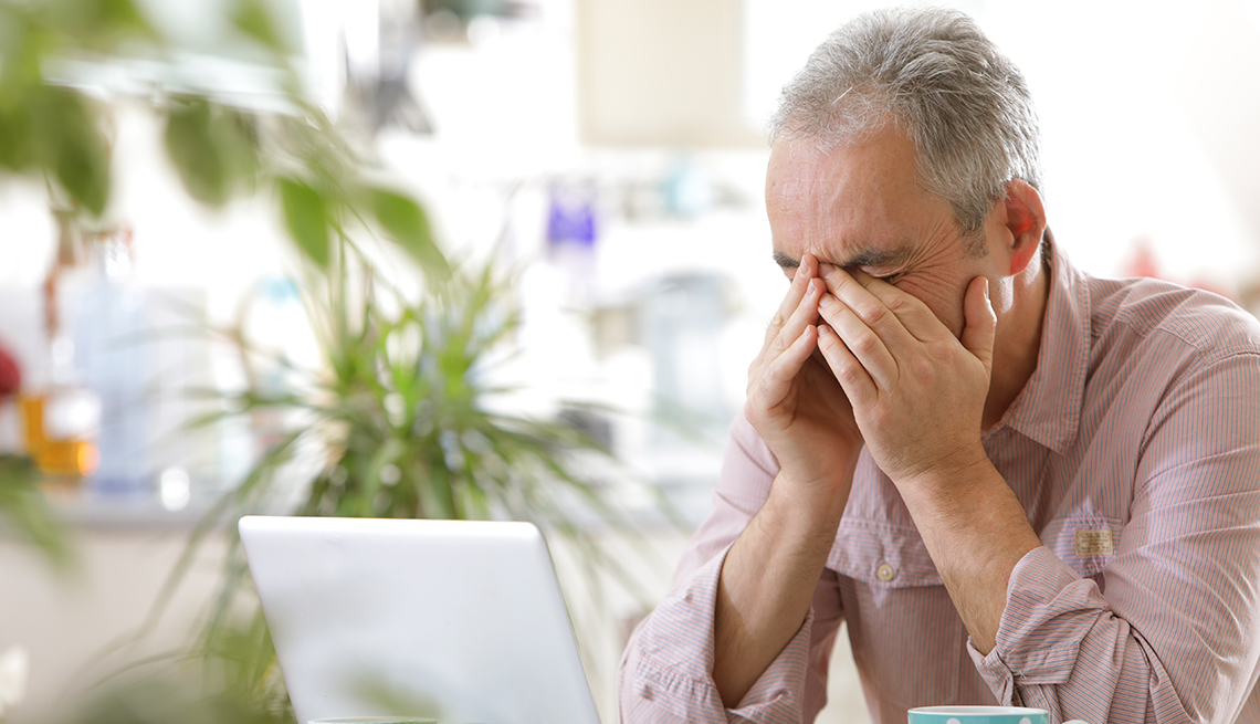 A man is sitting at a table with his head in his hands next to his laptop